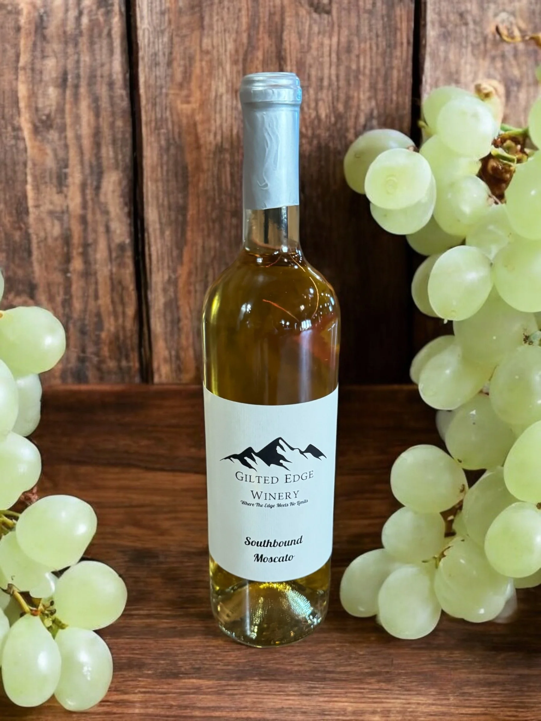 A bottle of white wine from Gilt Edge Winery, surrounded by green grapes on both sides, placed on a wooden surface against a wooden background.