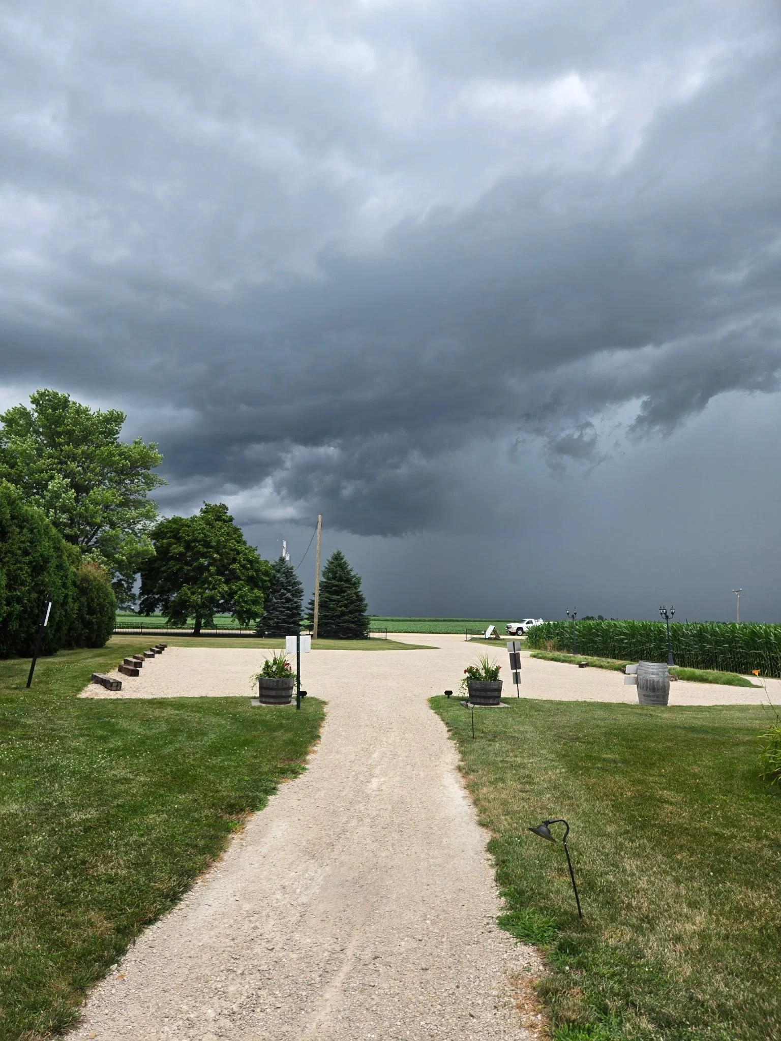 A gravel pathway in a rural area leads towards open fields, with trees and a cloudy dark sky overhead, indicating an approaching storm.
