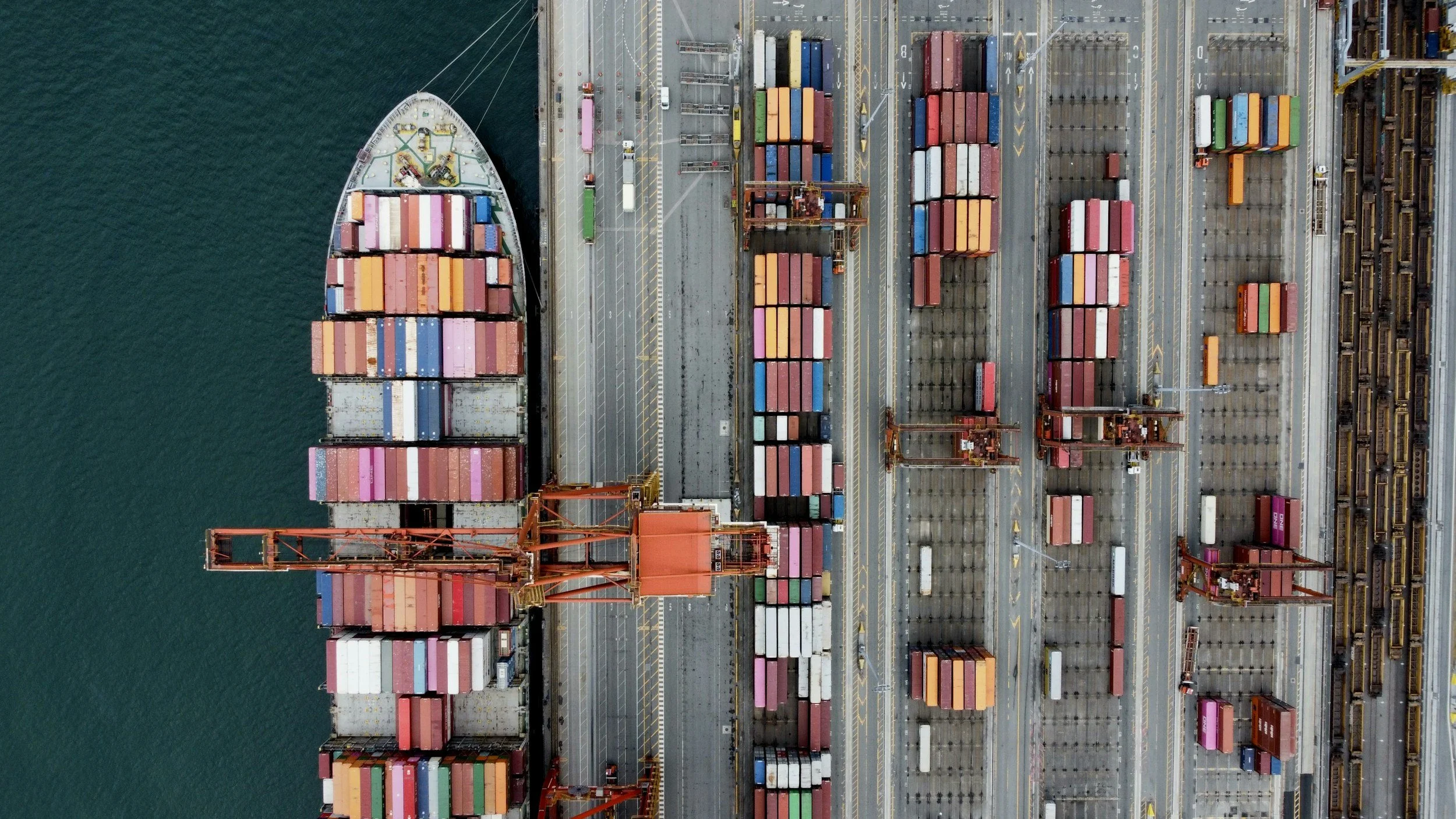 An aerial view of a shipping port with numerous multicolored shipping containers and large orange cranes, adjacent to a body of water.
