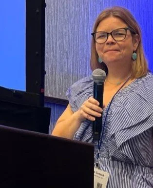A woman with glasses and earrings holding a microphone, standing behind a podium in front of a blue and gray background.
