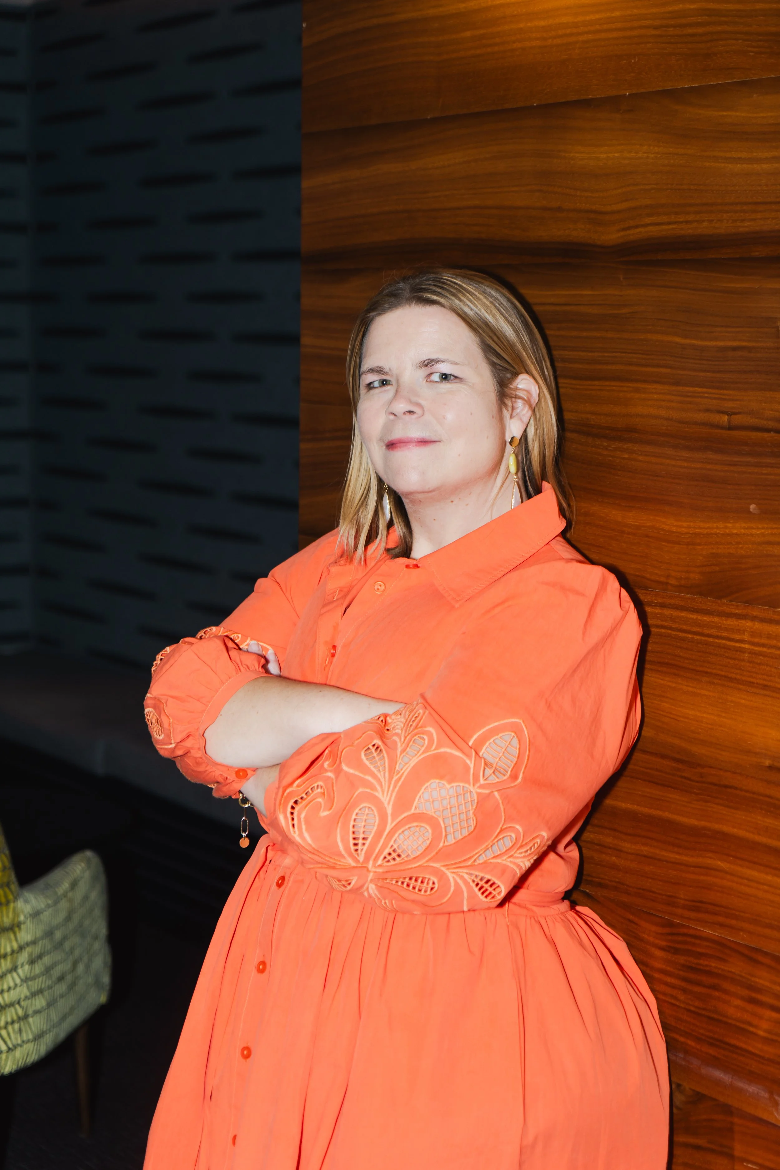 a woman with shoulder-length brown hair, wearing a bright orange dress with embroidered details on the sleeves, earrings, and crossed arms, standing against a wooden wall with a black panel on the left side.