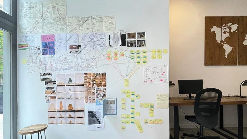 Whiteboard wall with interconnected notes, photos, and images in a workspace, with a nearby desk and chair, and a wooden wall art piece depicting a world map.