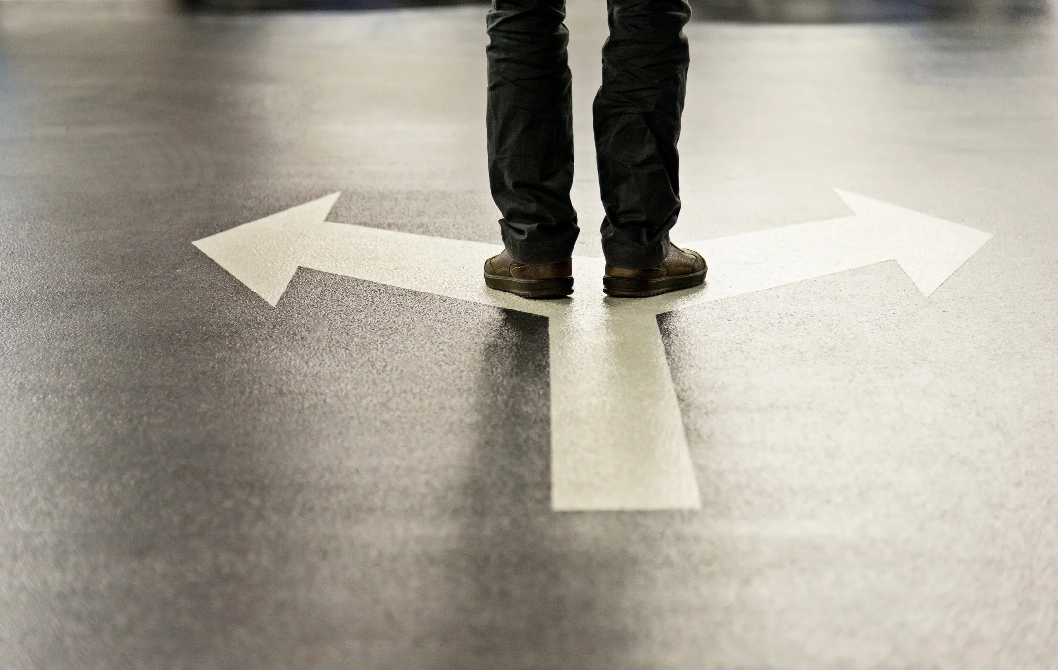 Person standing on a directional arrow painted on the floor, wearing black pants and brown shoes, in an indoor space.