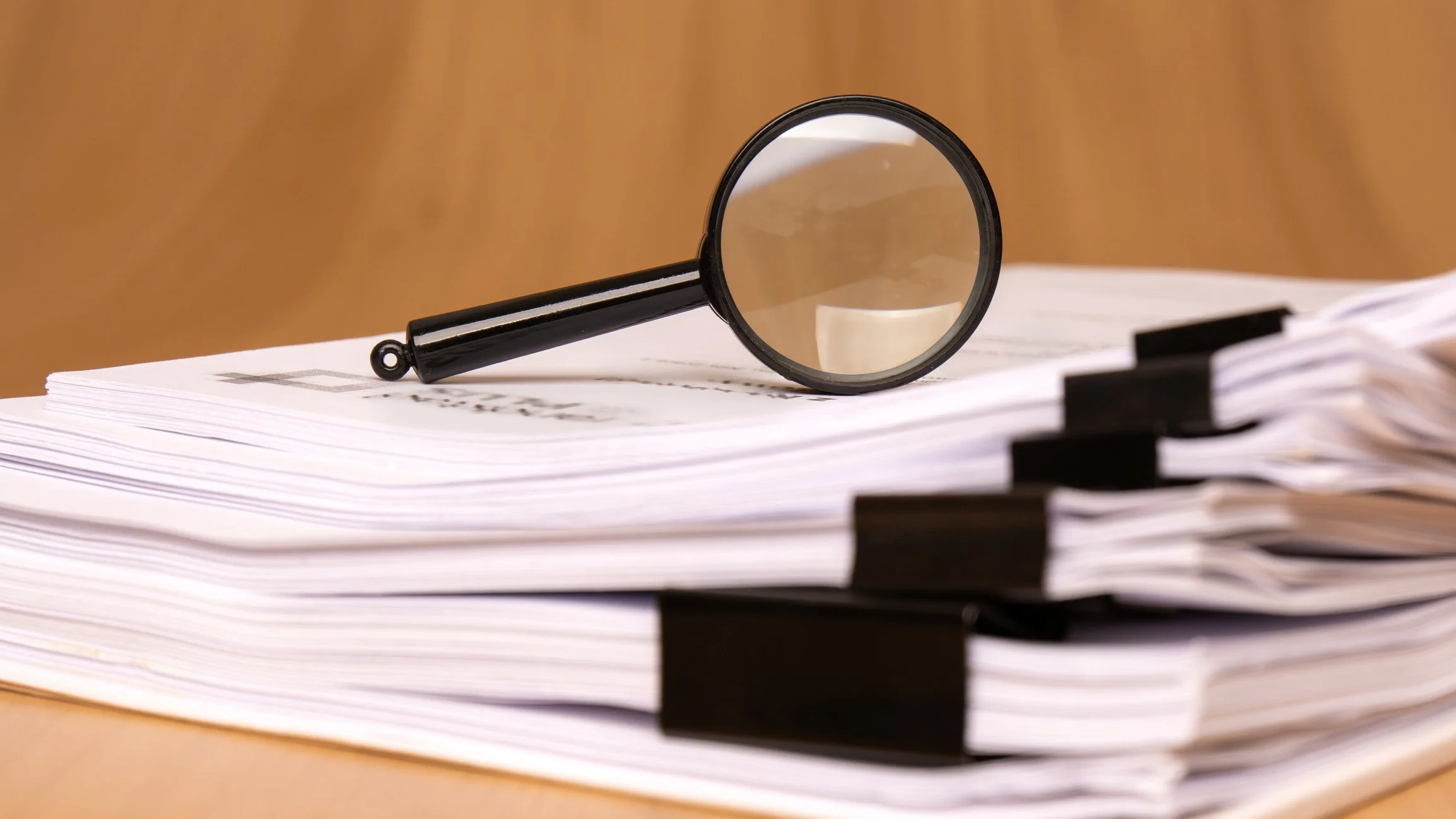 A black magnifying glass resting on a stack of paper files with black file tabs, on a wooden surface.