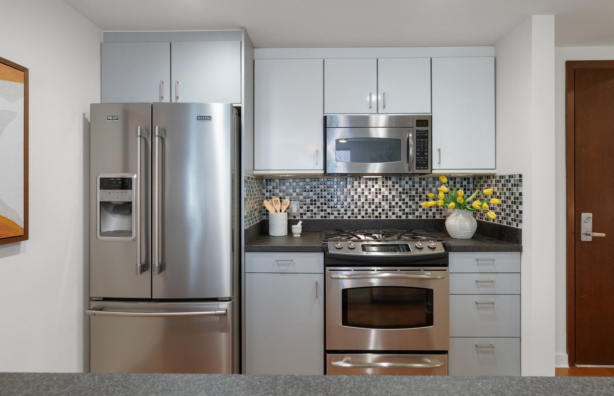 Modern kitchen with white cabinets, black countertops, stainless steel appliances, a mosaic tile backsplash, a vase with yellow tulips, and a built-in microwave, stove, and refrigerator.