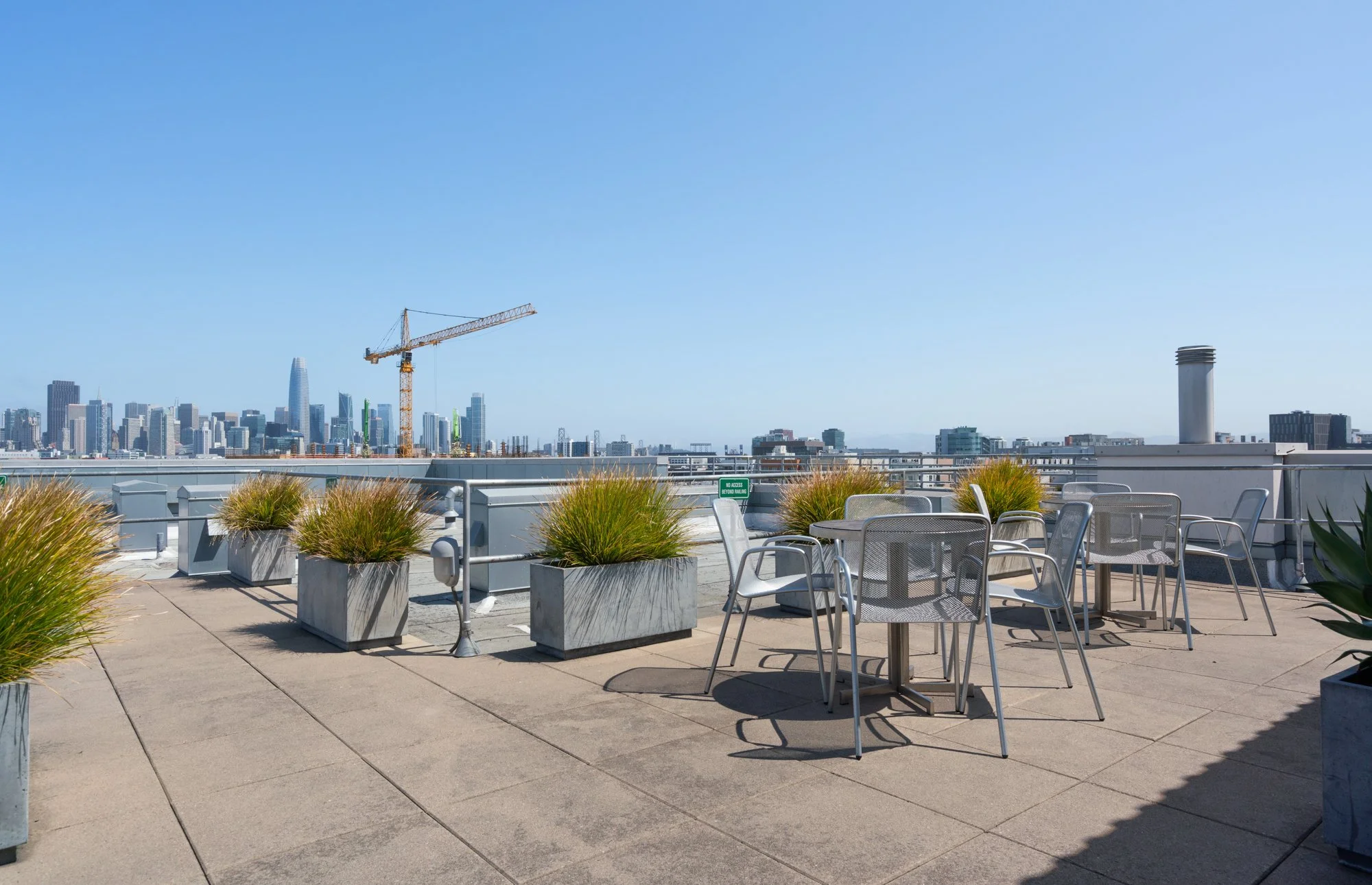 Rooftop terrace with round tables and metal chairs, large planters with grass, city skyline with tall buildings, construction crane visible in background, clear blue sky.