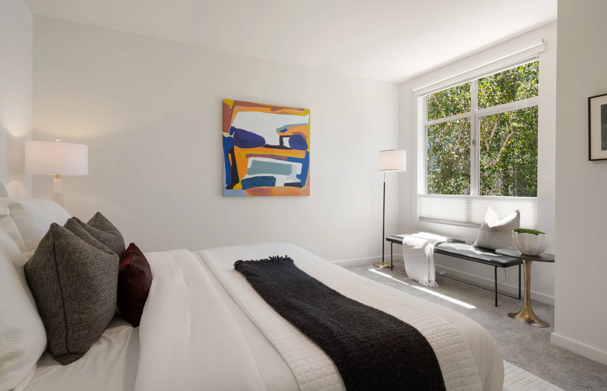 Bright bedroom with white walls, abstract colorful painting, large window with greenery outside, white upholstered bed with dark pillows, bedside lamps, bench with white towel and bowl, and a small side table.