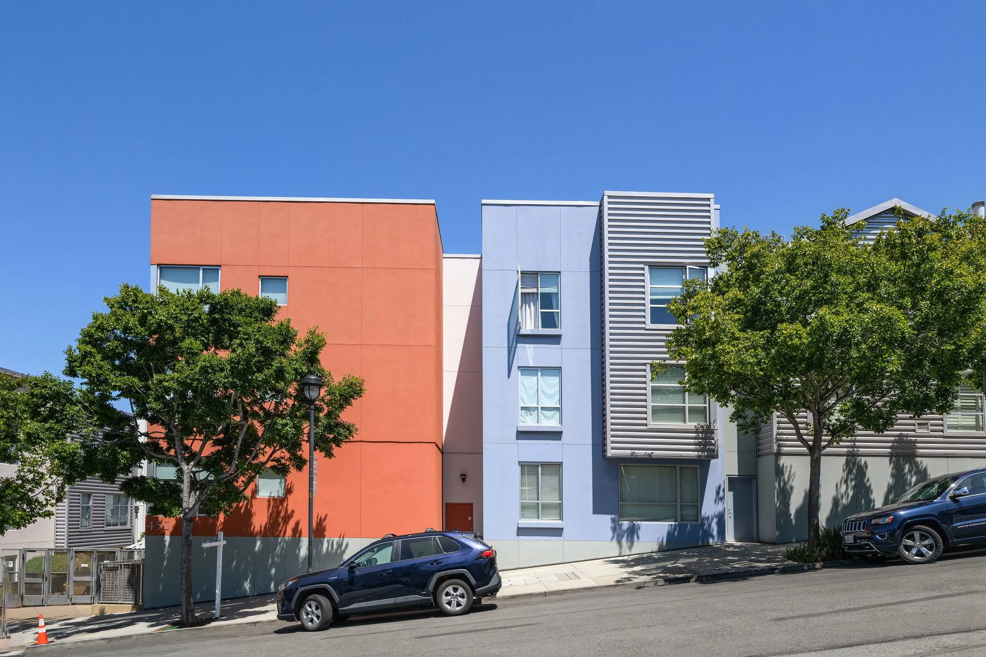 Colorful modern apartment building with orange, blue, and gray sections, green trees in front, parked cars on the street, blue sky overhead.