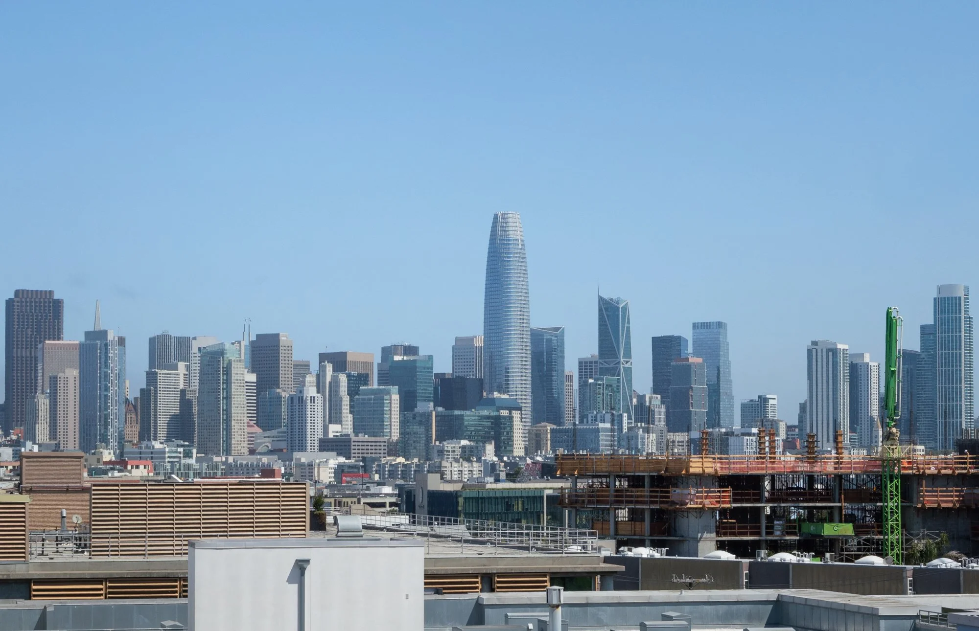 City skyline with tall skyscrapers under a clear blue sky, including a prominent cylindrical building and a construction site in the foreground.