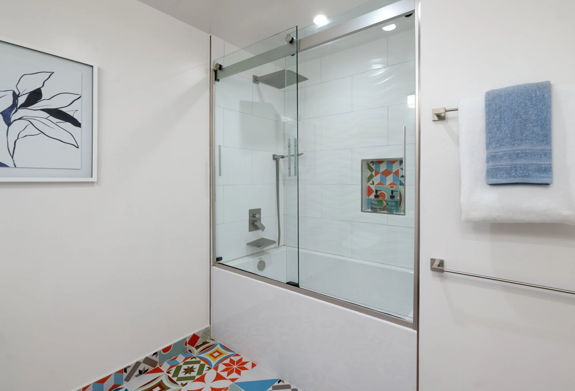 A modern bathroom with a glass-enclosed shower, colorful patterned floor tiles, a towel rack with blue towels, and a framed leaf artwork on the white wall.