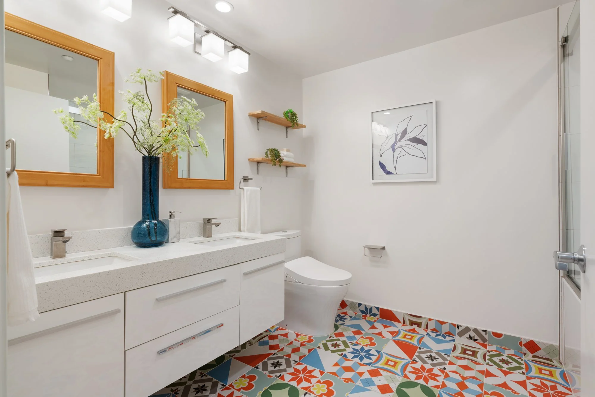 Modern bathroom with white vanity, two wooden framed mirrors, decorative tile floor, and wall art.