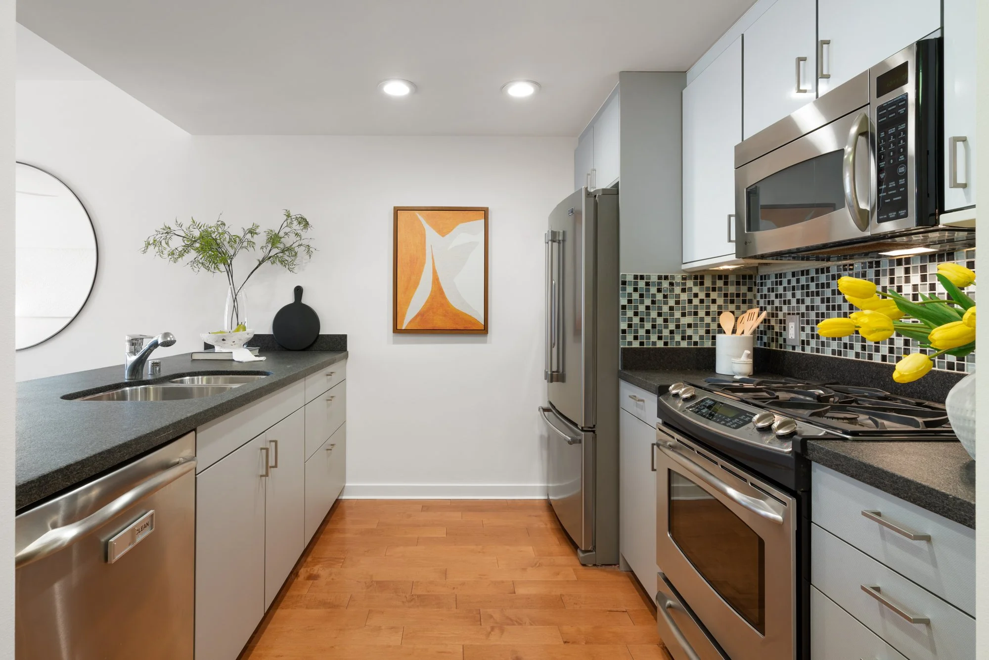 Modern kitchen with white cabinets, stainless steel appliances including a refrigerator, microwave, and stove, black granite countertops, a mosaic tile backsplash, yellow tulips in a white vase, and a decorative black circular object on the wall.