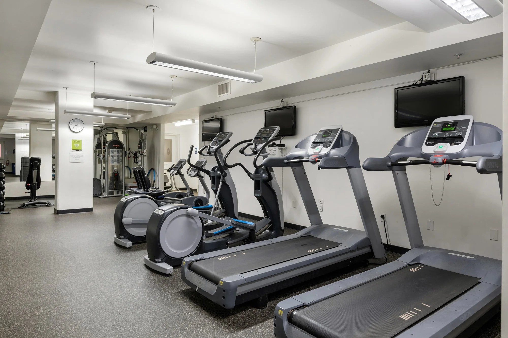 A gym room with exercise equipment including treadmills, ellipticals, and television screens on the wall.