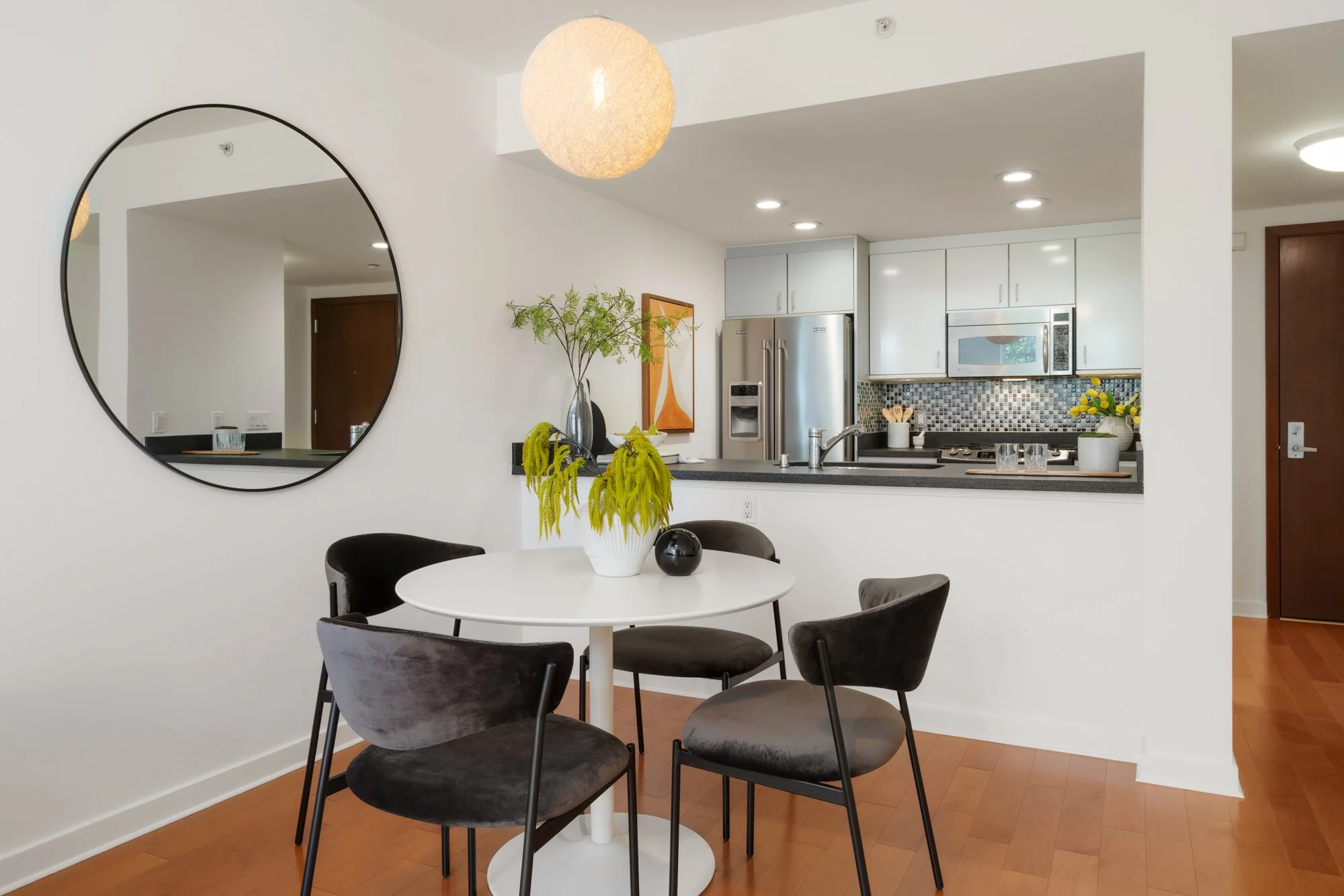A dining area with a round white table surrounded by five black velvet chairs. A vase with green plants and a small black decorative item are on the table. In the background, part of a kitchen with white cabinets, a stainless steel refrigerator, a microwave, and a tiled backsplash are visible. A large circular mirror hangs on the white wall, and warm lighting is present.