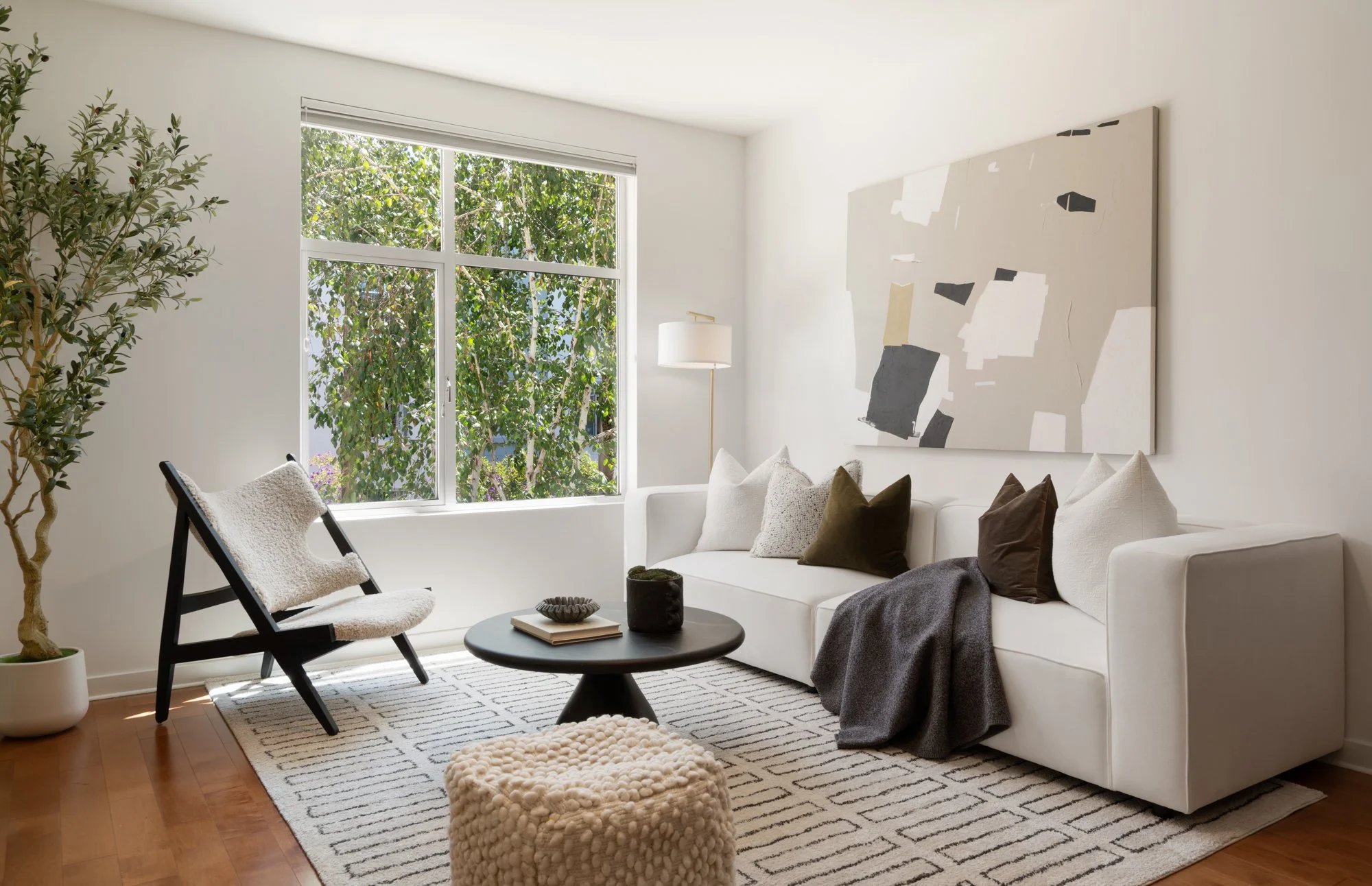 A living room with white walls, a large window showing green trees outside, a white sofa with multiple pillows, a black coffee table with books and decorative items, a beige plush ottoman, a black armchair with white cushion, a tall floor lamp, a lar