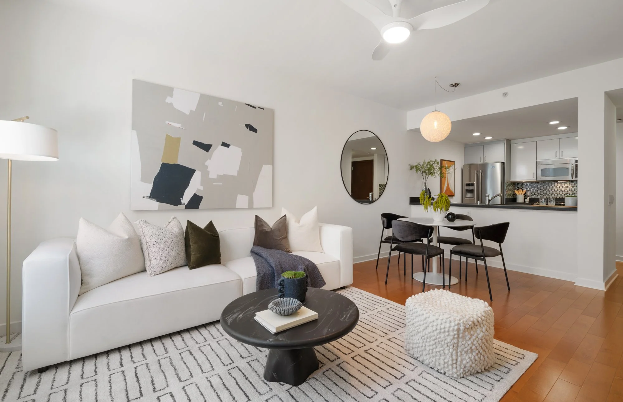 Modern living room with white sofa, abstract wall art, black round coffee table with books, beige and black pillows, a white textured ottoman, a black dining table with black chairs, kitchen in background with stainless steel fridge, microwave, and c
