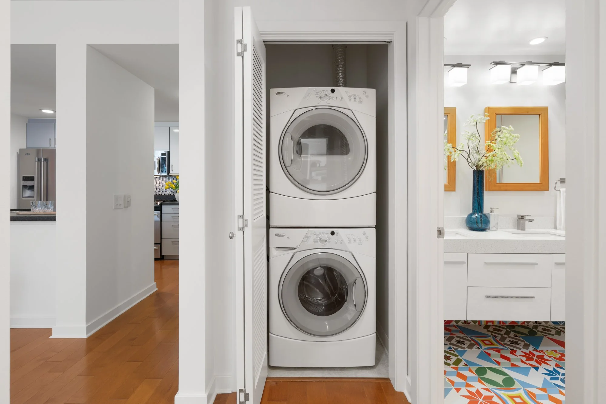 A stacked washer and dryer inside a closet with white louvered doors, adjacent to a bathroom with a blue vase and a mirror, in a modern home.