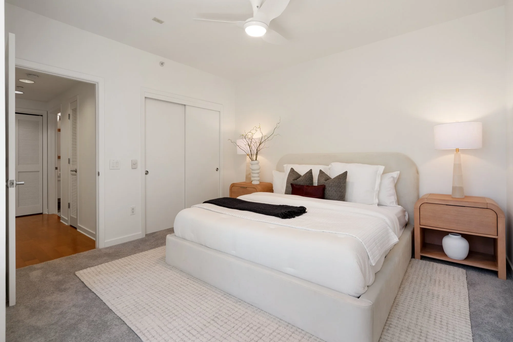 A modern bedroom with a white bed, decorative pillows, a bedside table with a vase and a lamp, and a ceiling fan with lights. The walls are white, and there's a wooden floor and a light-colored rug.