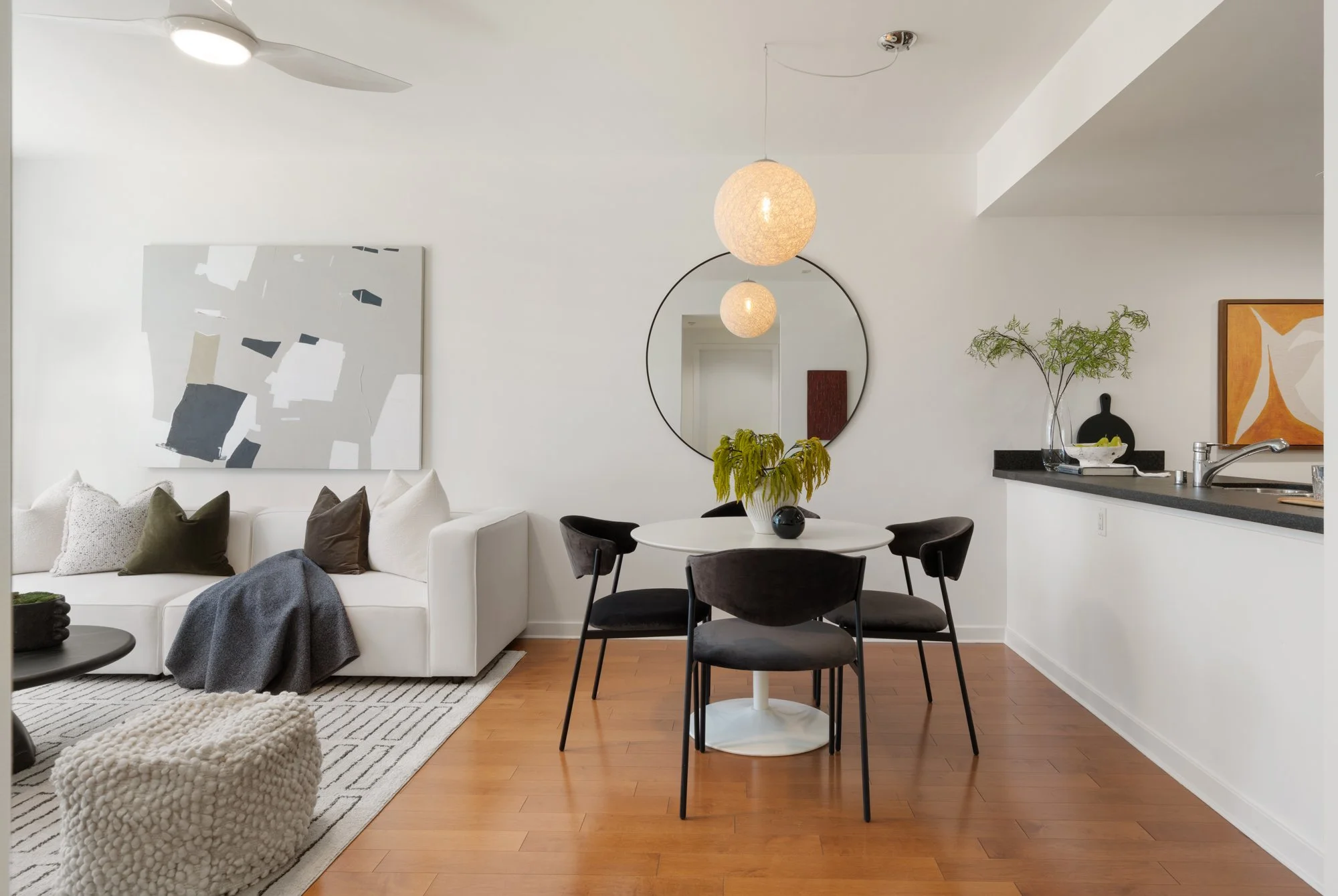 Living room and dining area with white walls and ceiling, wooden flooring, white sofa with multiple pillows, black circular table, abstract wall art, round mirror, hanging light fixtures, plants, and kitchen counter with a faucet.