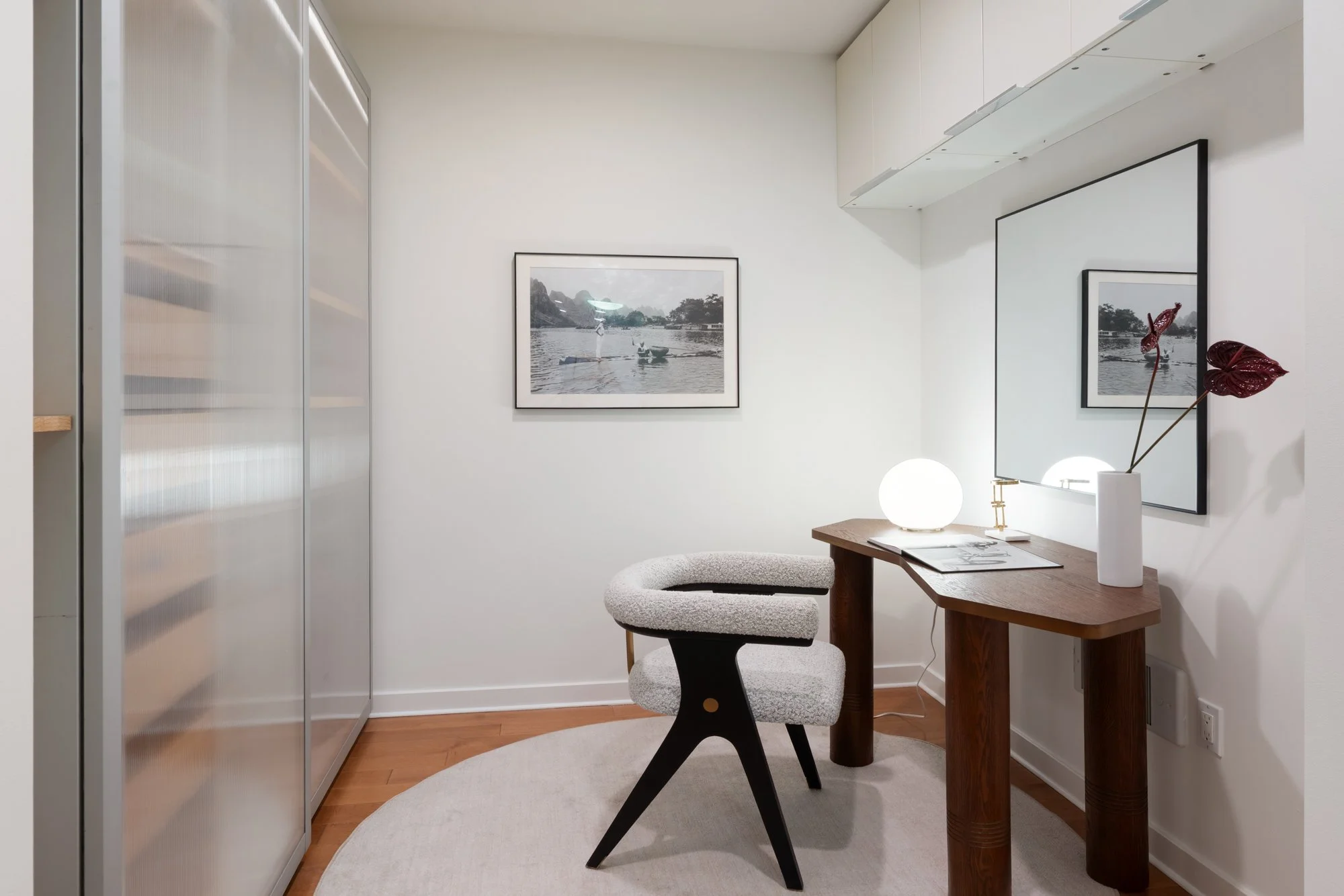 A modern minimalist workspace with a wooden desk, a white upholstered chair, a large mirror, framed black and white photographs of river scenes on the wall, a white vase with decorative red leaves, a spherical table lamp, and light-colored wood floor