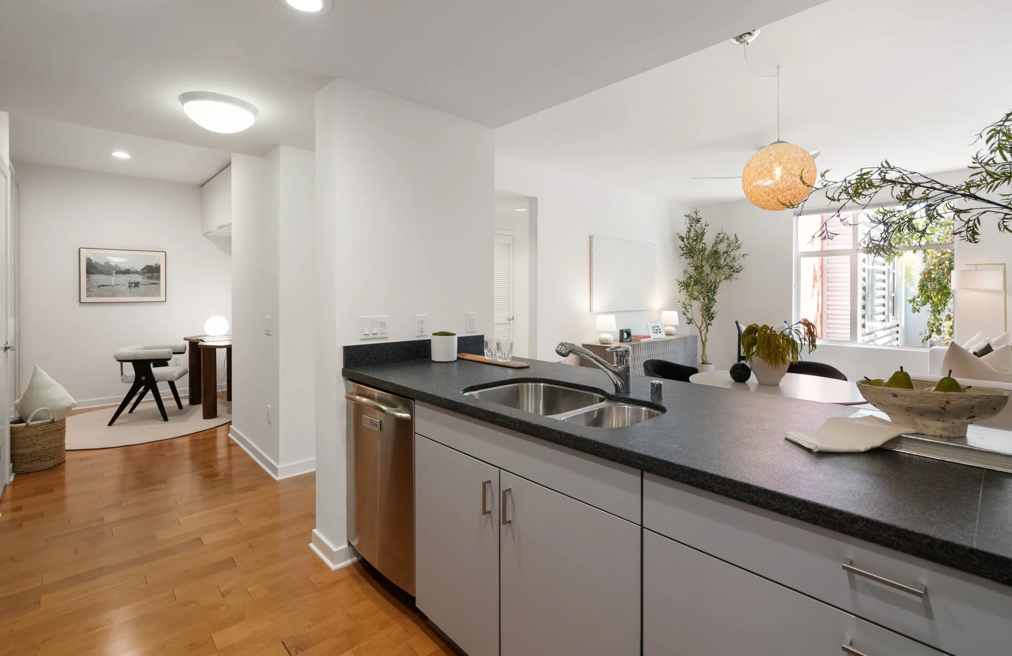 Open-concept kitchen and living room with white walls, hardwood flooring, modern lighting, window with outdoor plants, kitchen island with sink, and living area with furniture and decor.