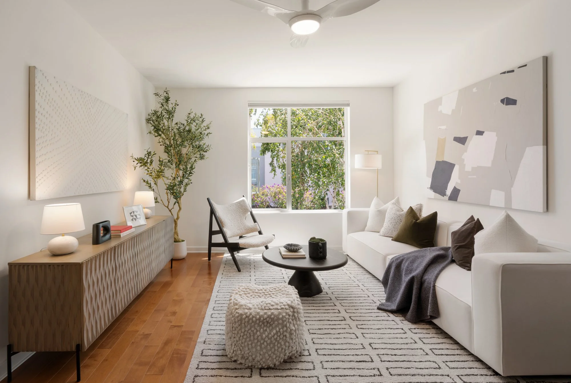 Bright living room with large window, white sofa with brown and white pillows, black coffee table, patterned rug, comfortable chair, sideboard with lamps and books, and artwork on white walls, with a ceiling fan.