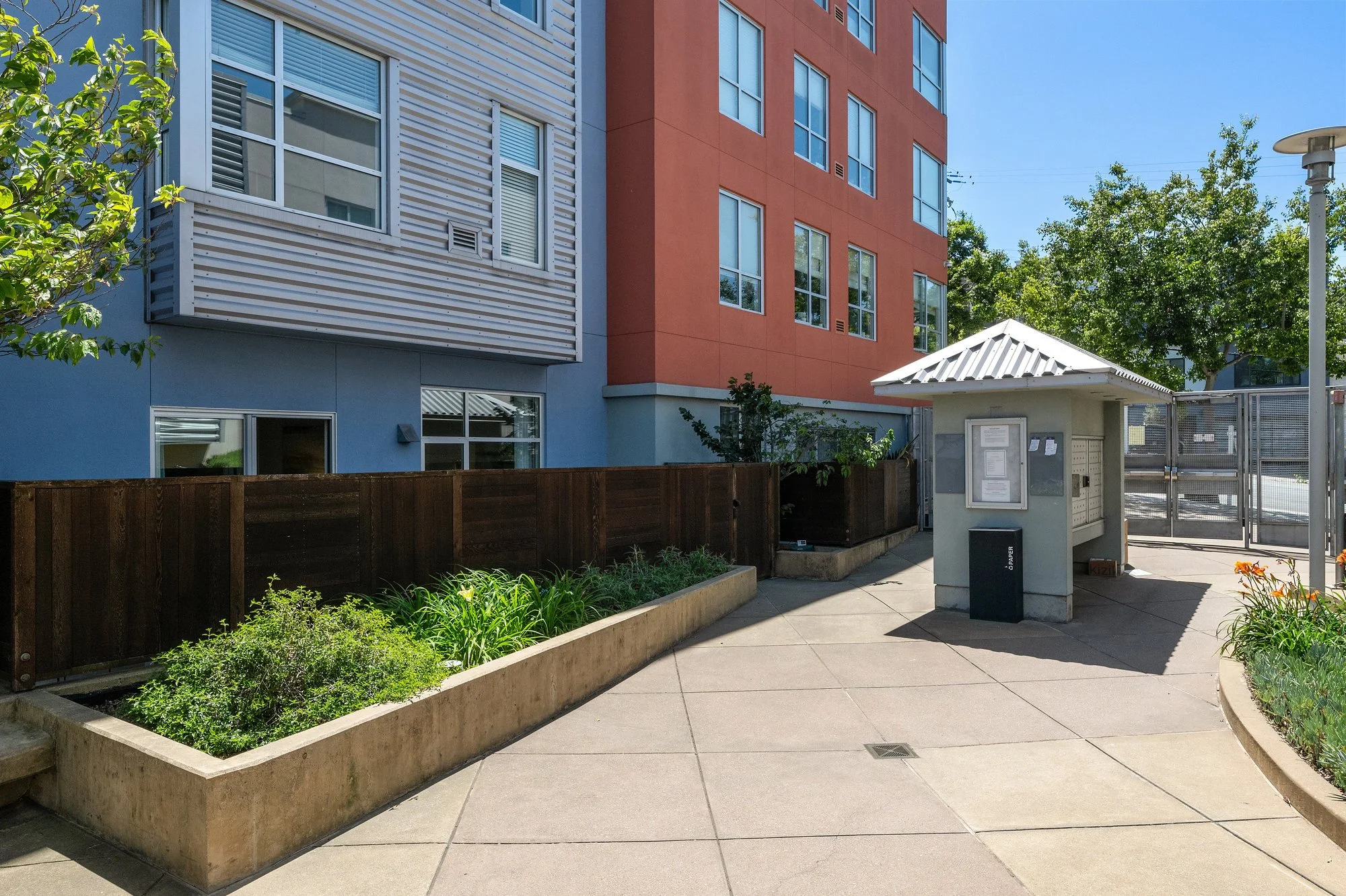 Exterior view of a modern multi-story apartment building with blue and red walls, surrounding trees, a covered mailbox area, and a sidewalk with garden beds