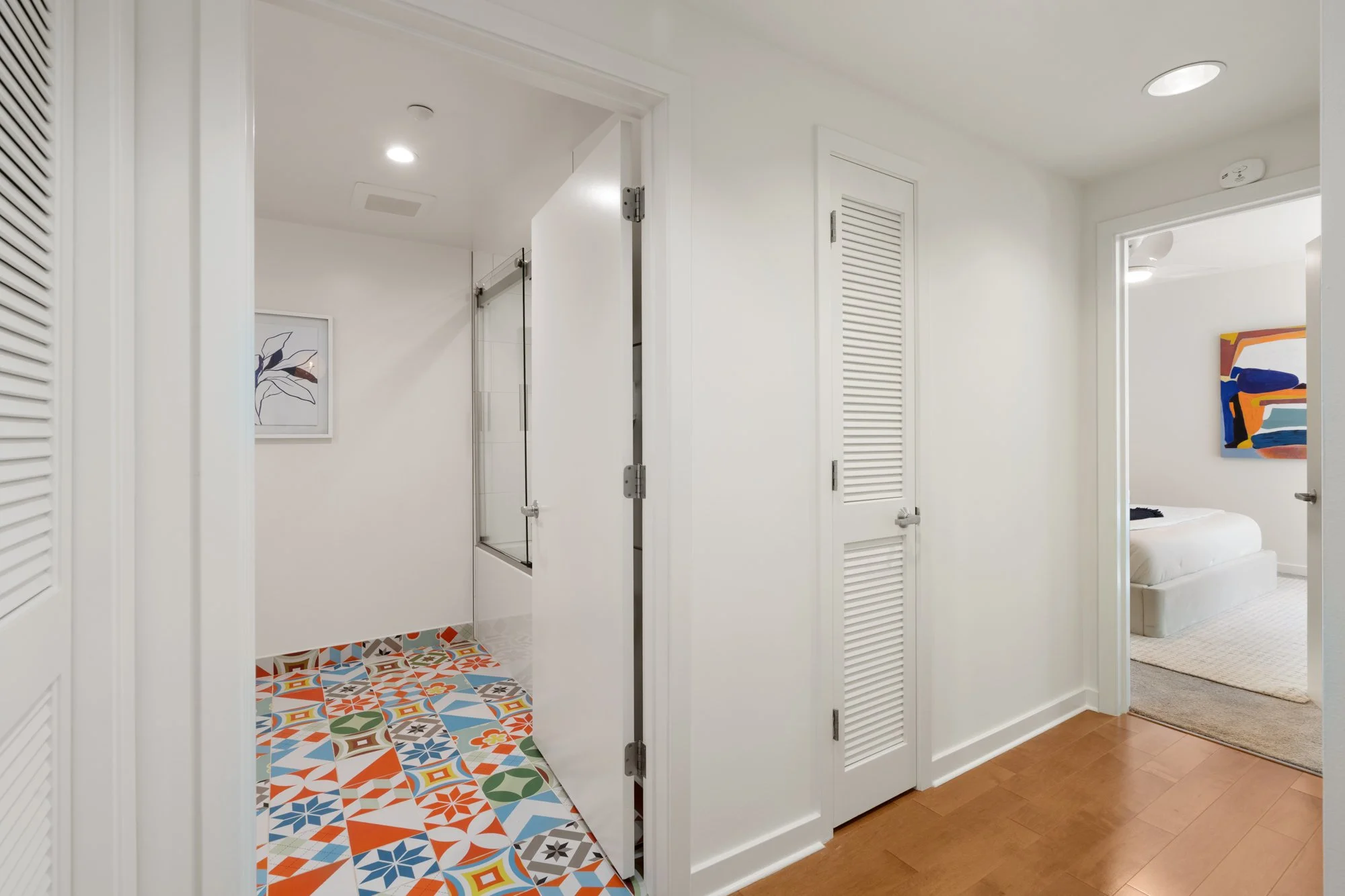 View of a modern bathroom with a glimpse into a bedroom. The bathroom has white walls, colorful patterned floor tiles, and a glass shower enclosure. The door to the bedroom is open, showing an abstract colorful painting and a white bed.