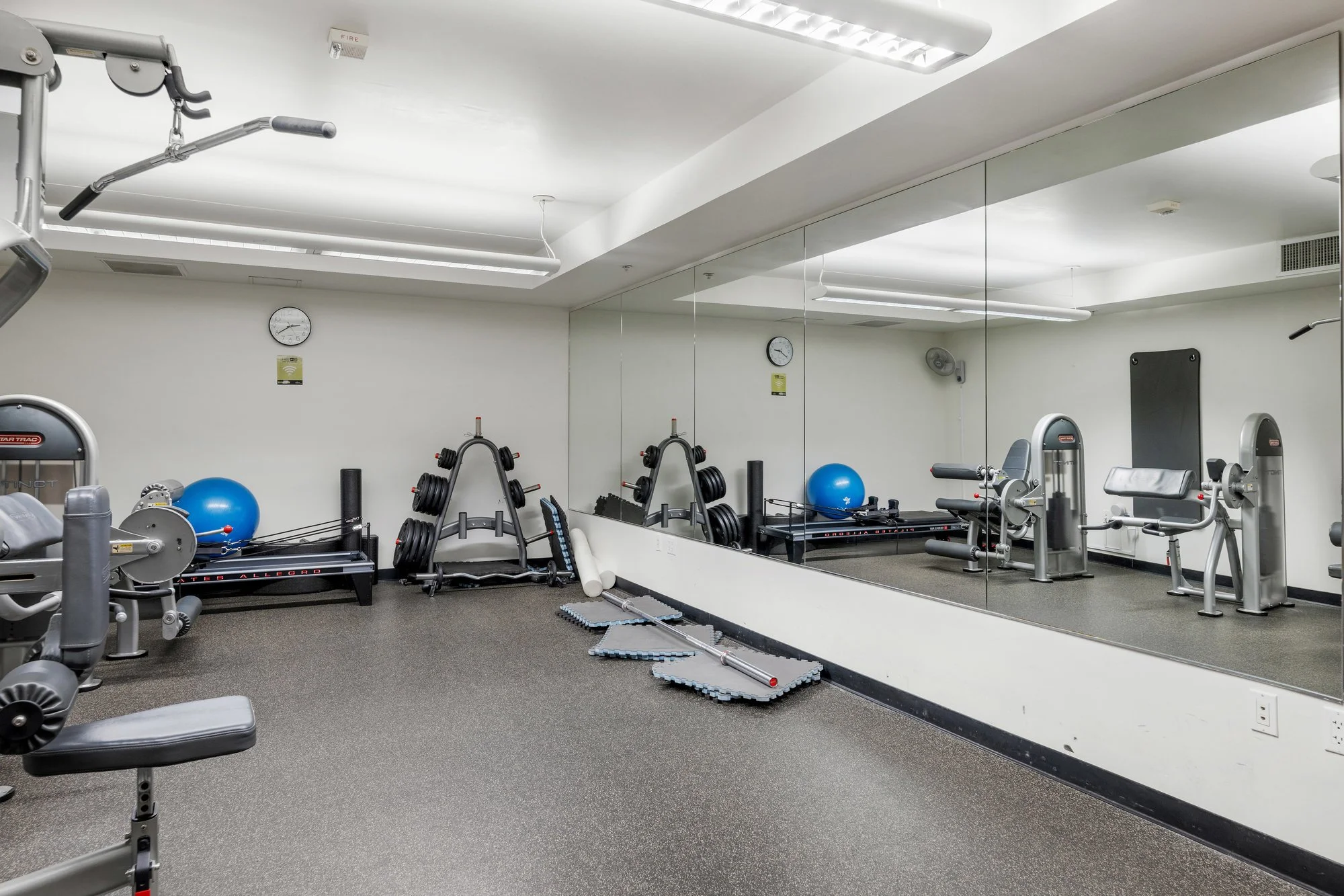Interior of a gym with various workout machines, weights, exercise balls, and mats. Large mirror on the wall reflects the room. Ceiling has bright fluorescent lights, and there is a clock on the wall.