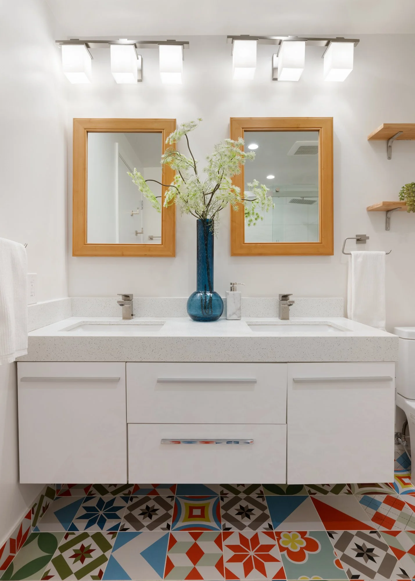 Bathroom with two mirrors with wooden frames, white vanity with storage drawers, a blue vase with white and green flowers, a marble soap dispenser, colorful patterned floor tiles, and bright overhead lights.