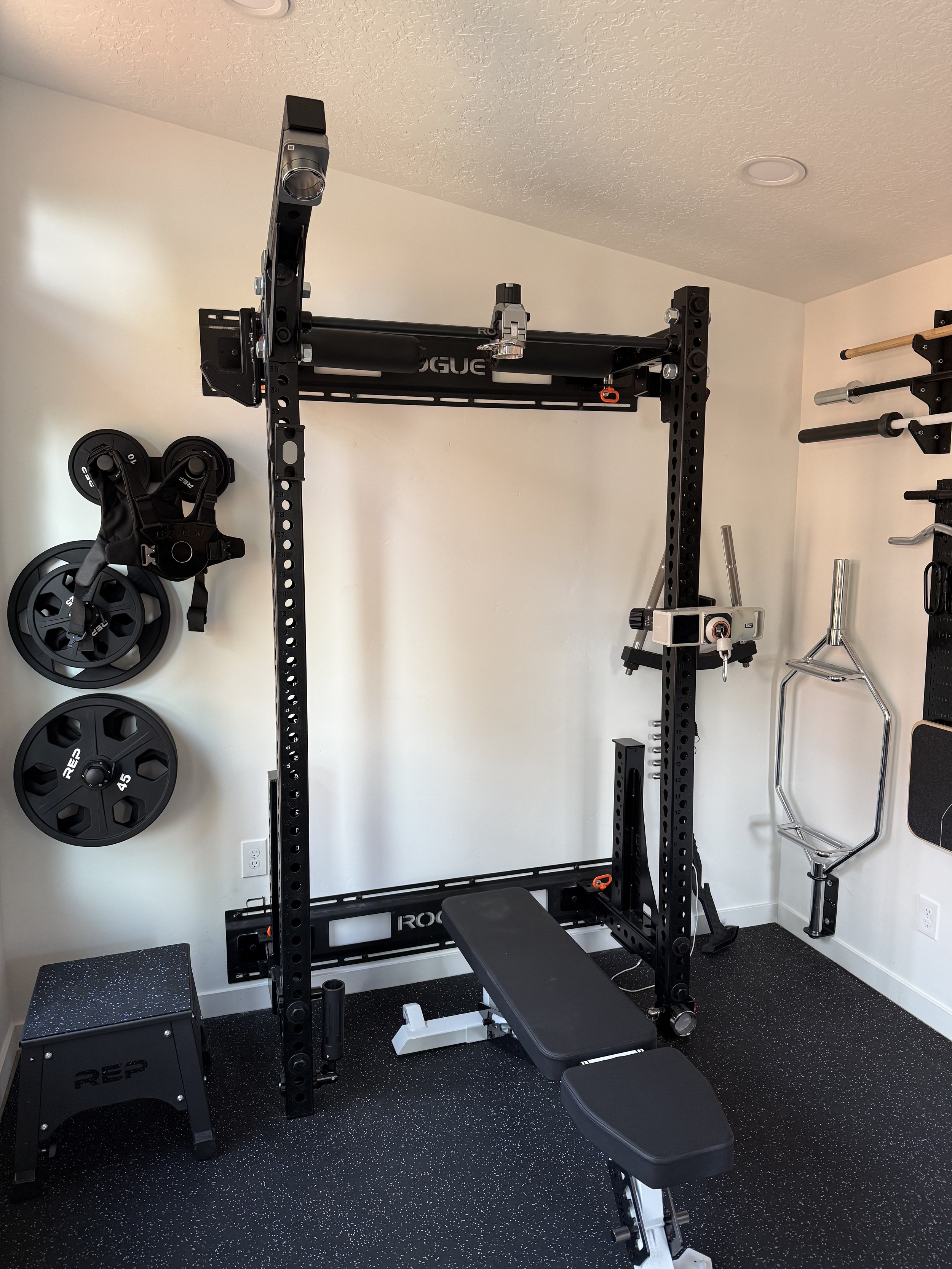 A home gym with a power rack, adjustable bench, weight plates, and various workout equipment on a black speckled rubber floor.