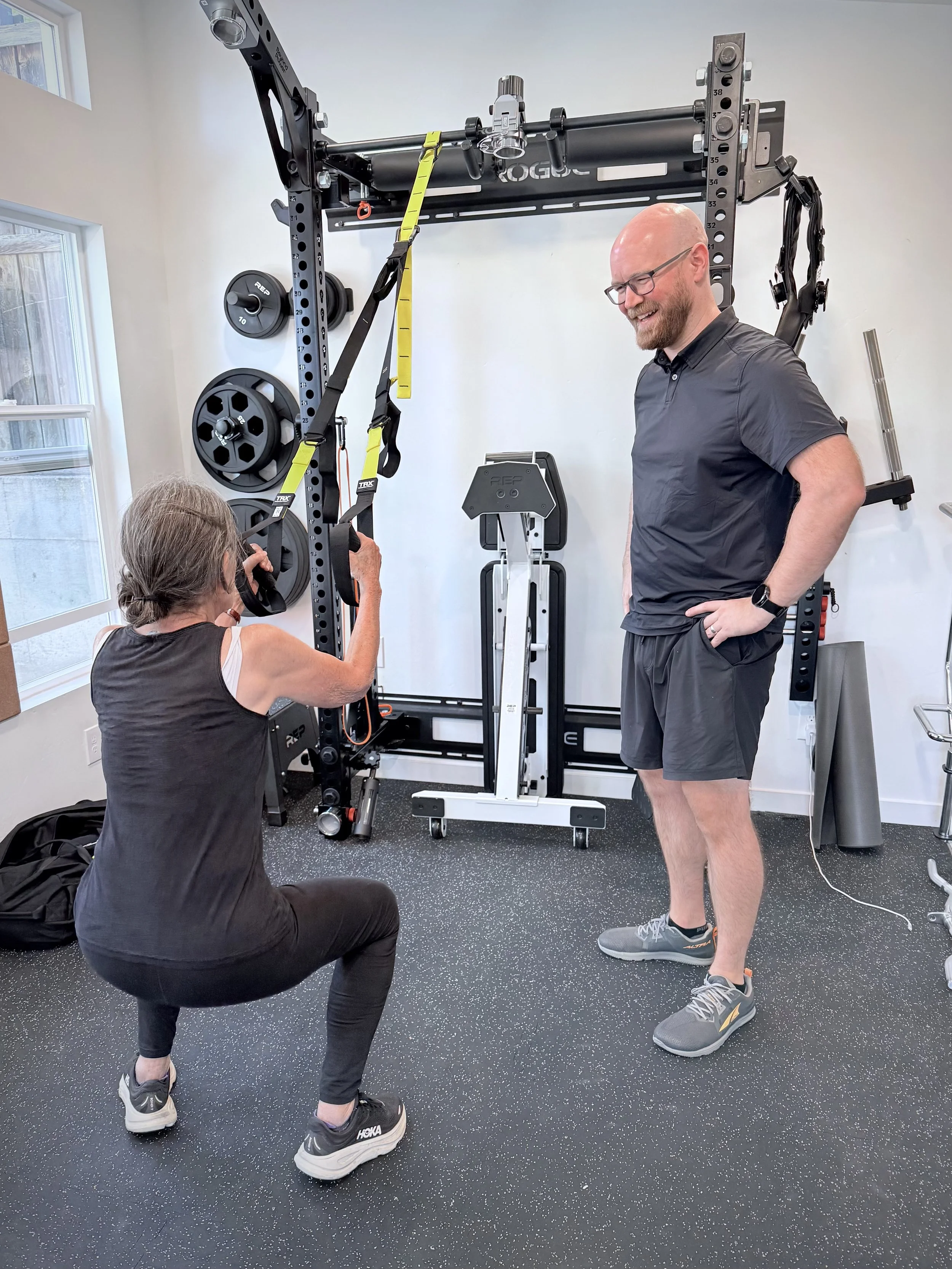 A woman doing a squat exercise with a suspension trainer, while a man in athletic clothes watches and smiles in a gym with workout equipment.