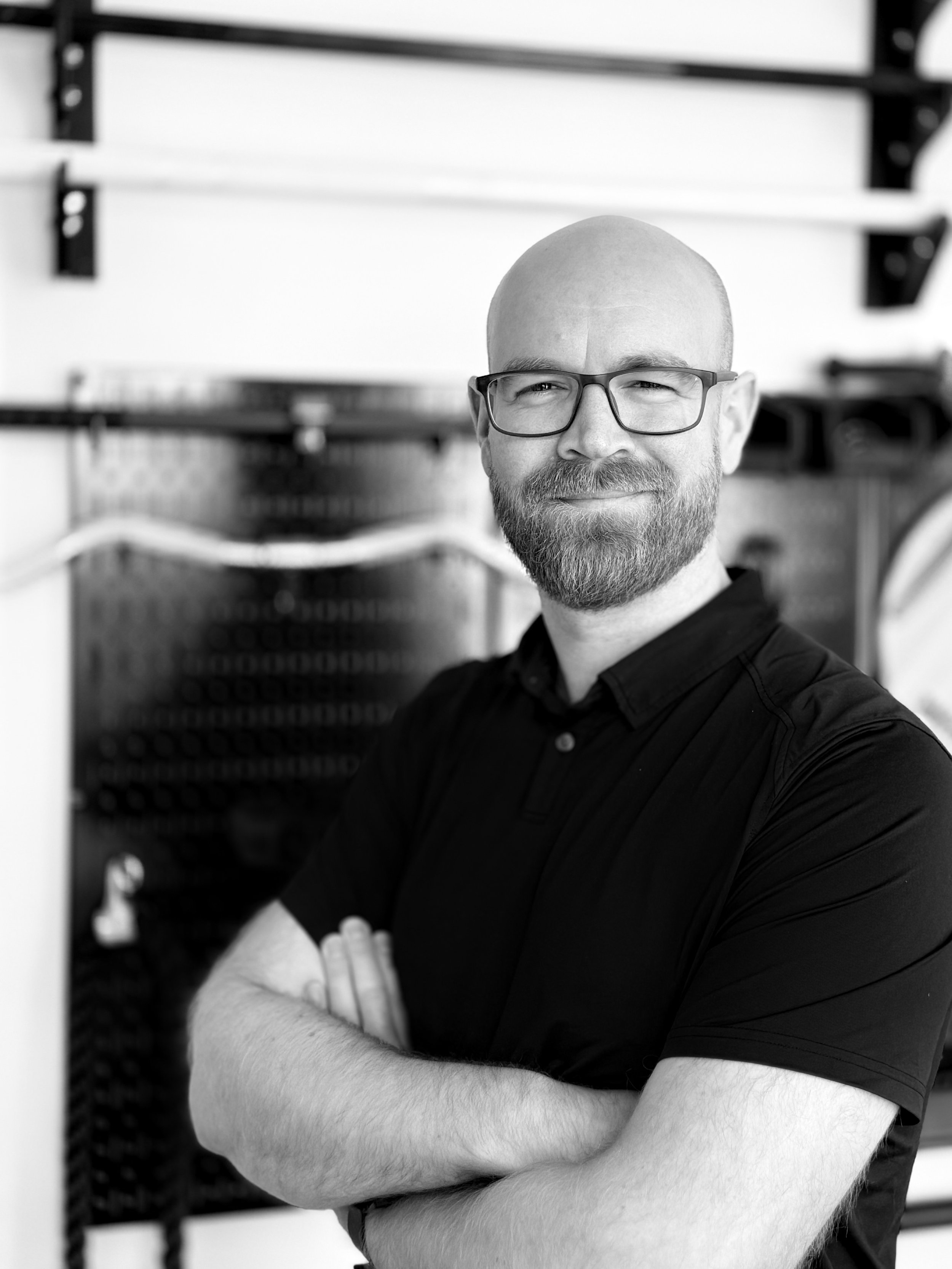 A black and white photo of a bald man with glasses and a beard, smiling with folded arms, standing in front of gym equipment.