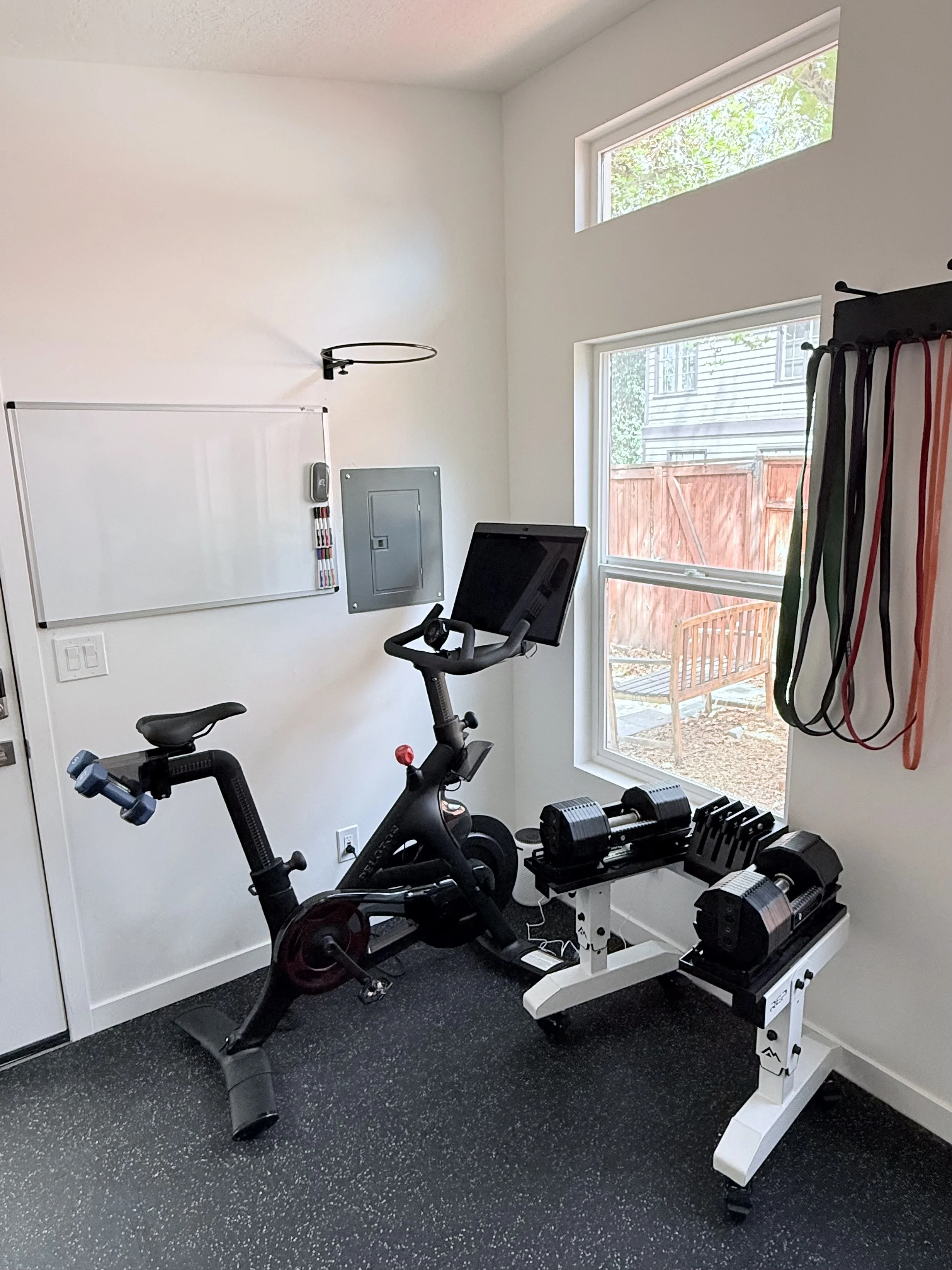 Home gym with exercise bike, dumbbells, resistance bands, and workout equipment in front of two windows and a whiteboard on the wall.