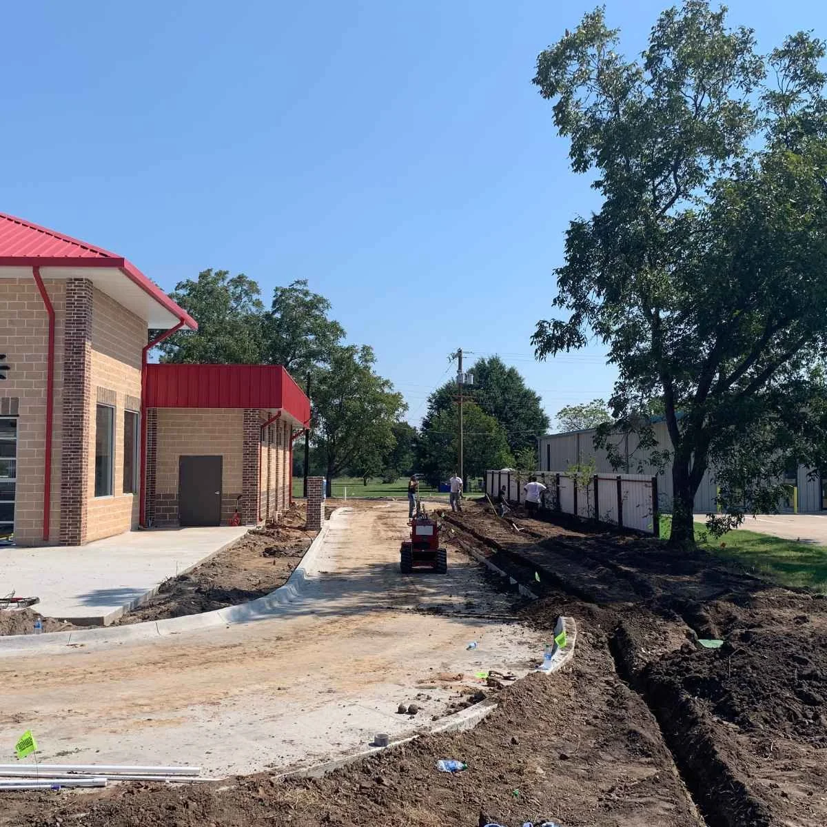 Construction site on a sunny day with a partially paved sidewalk, dirt, and construction workers during irrigation system installation. There are trees, a brick building with a red metal roof, and a white building in the background.