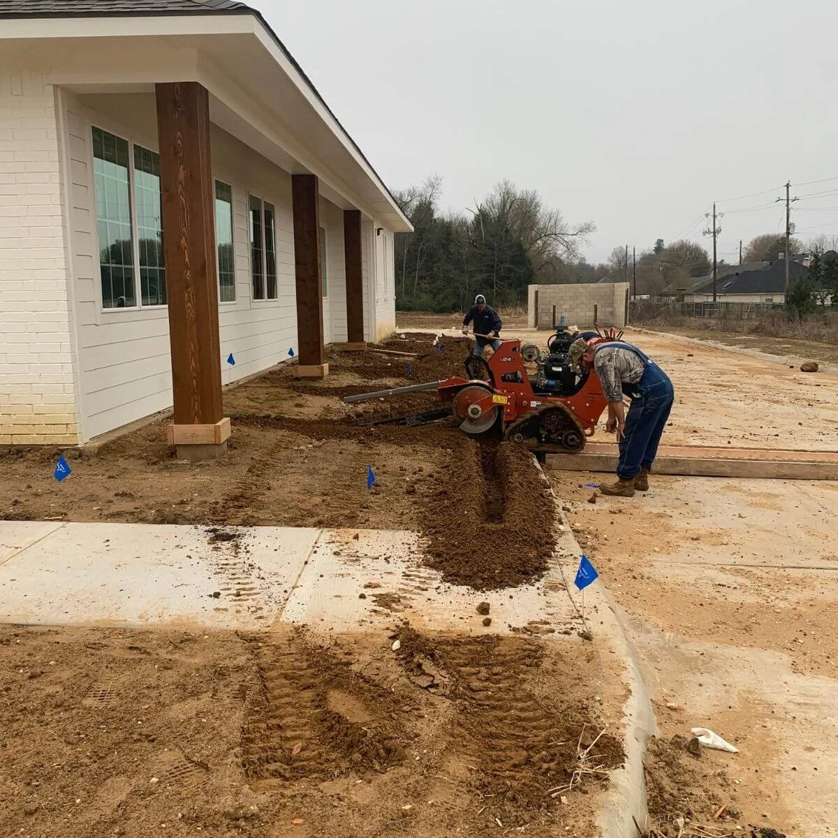 Construction workers leveling soil outside a building with a walk-behind tractor, preparing for irrigation system.