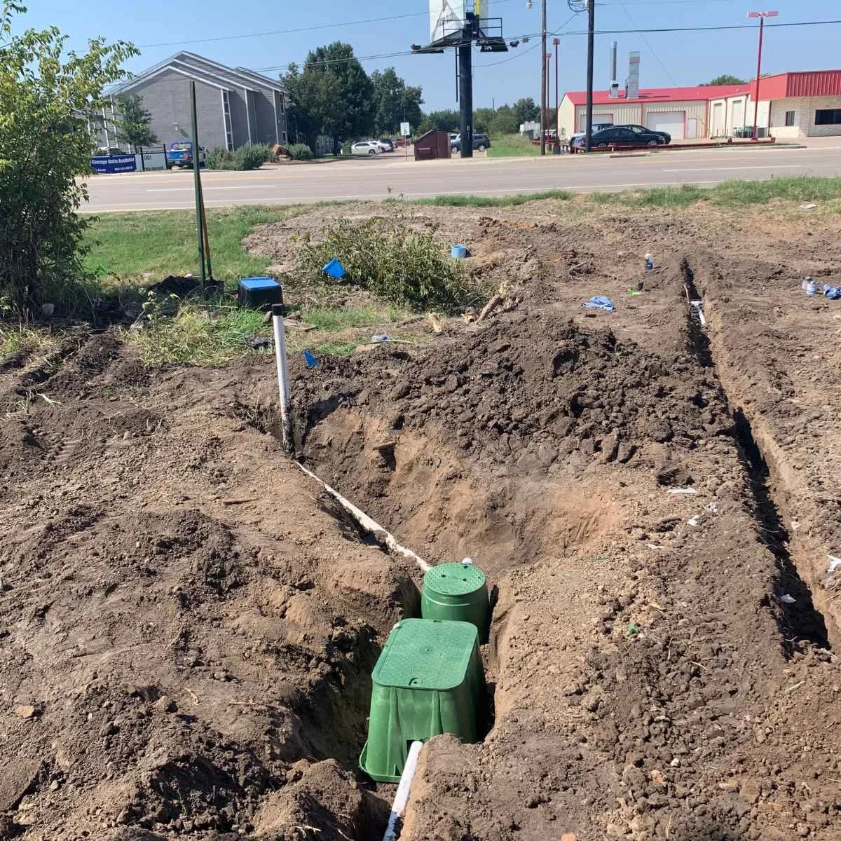Commercial site with trenches shown during irrigation system installation in front of a road with buildings and parked cars, under a sunny sky.
