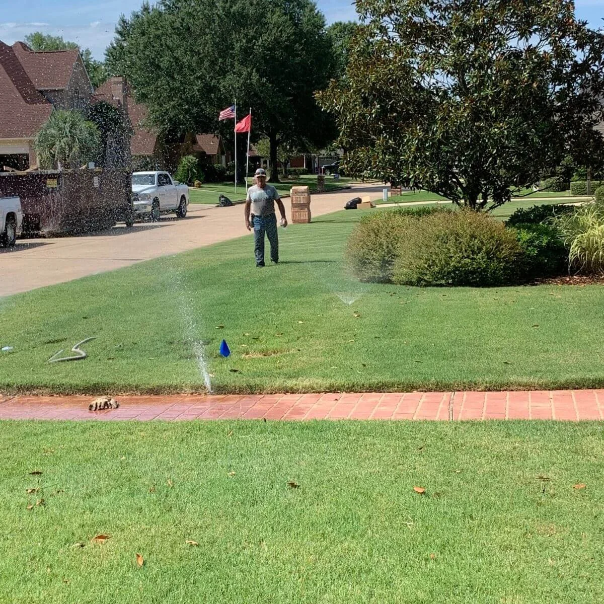 A man watering the lawn with a garden sprinkler on a sunny day in a suburban neighborhood with green grass, trees, and houses in the background.