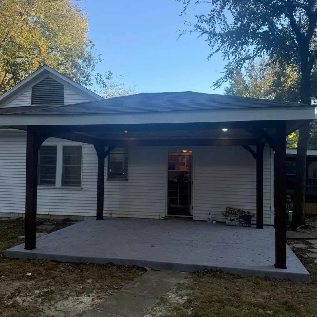 Backyard patio with a covered pergola, house with windows, and a door leading inside, surrounded by trees and a clear sky.