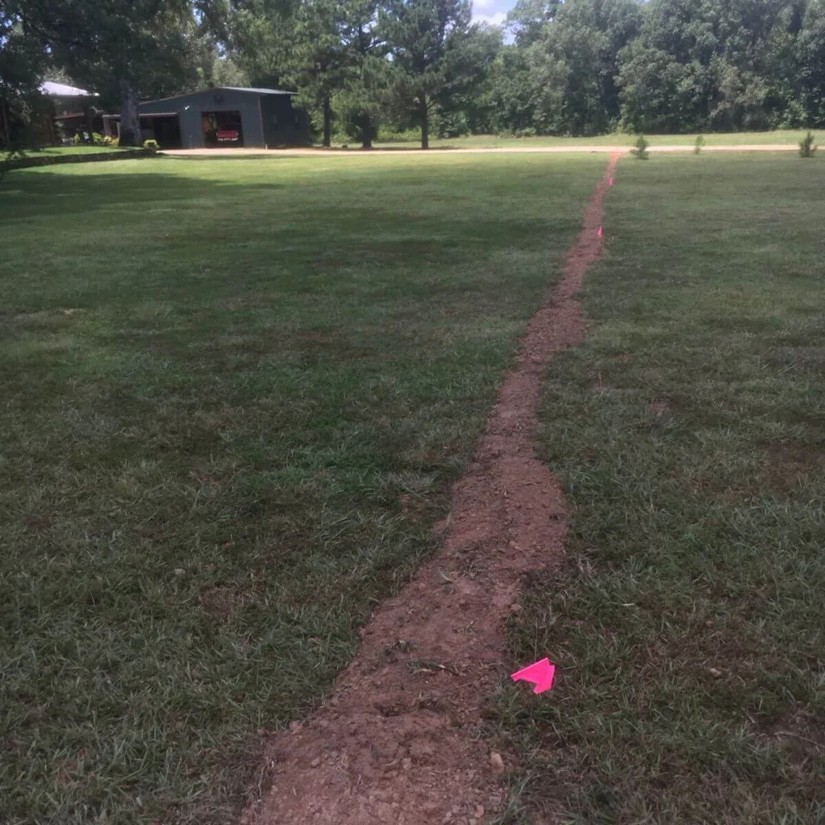 A dirt path marked with pink flags stretches across a green lawn towards a dark-colored building with trees in the background.