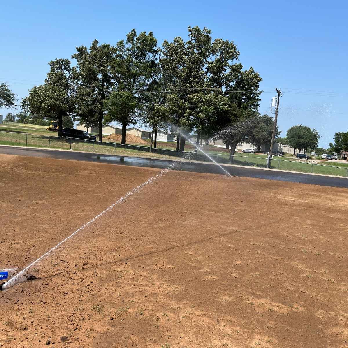 A baseball field being sprayed with newly installed sprinkler system. There is freshly watered dirt infield, green grass, and tall trees in the background under a clear blue sky.