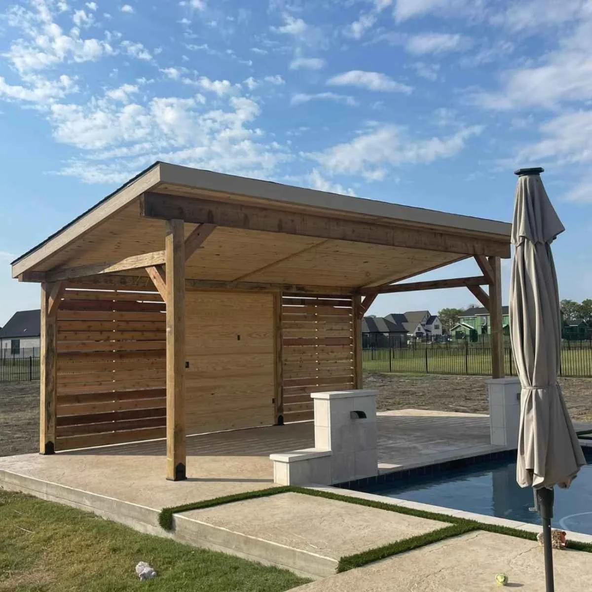 A backyard with a wooden patio addition over a concrete slab, adjacent to a swimming pool with a closed beige umbrella on the right side, and a lawn with a small patch of grass in the foreground. 