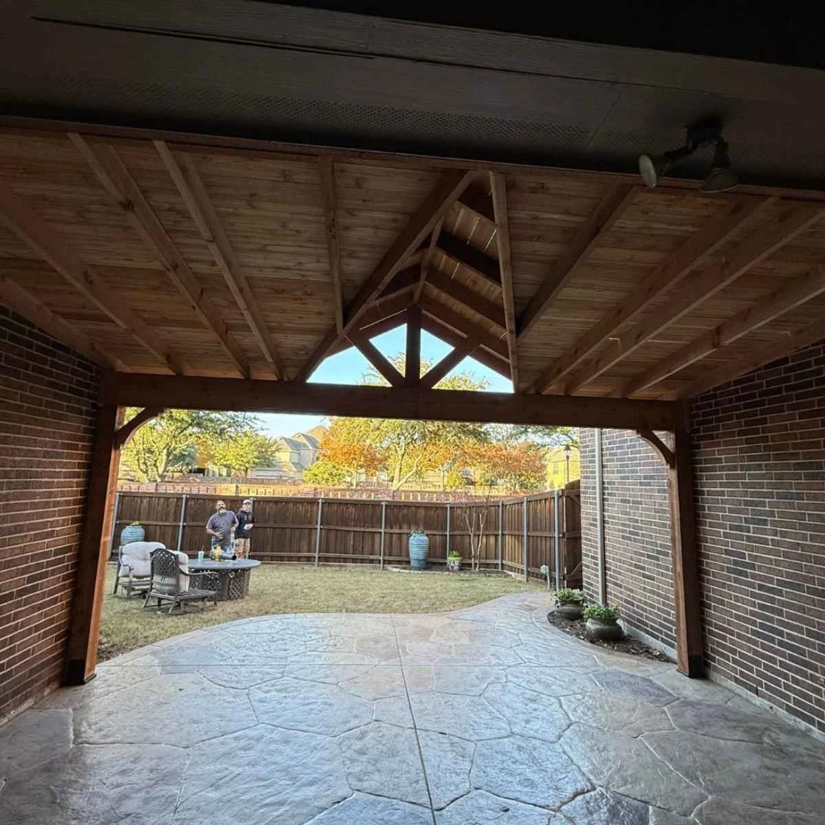 View of a backyard patio with a wooden roof structure, brick walls, and a concrete stamped floor. 