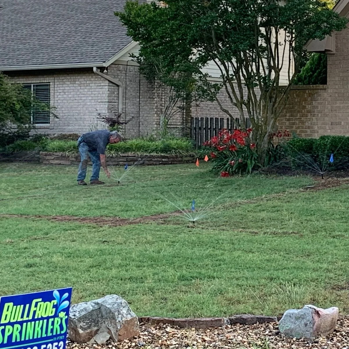 Sprinkler system shown in a residential yard with green grass, shrubs, flowers, and a large tree, with a house in the background in Paris, Texas