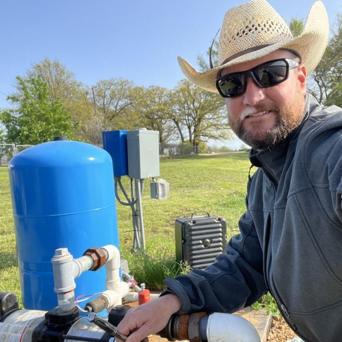 Bullfrog Sprinklers owner, Jeremiah Handlin wearing a cowboy hat, sunglasses, and a dark jacket smiling outdoors next to a blue water pressure tank and associated plumbing equipment in Paris, Texas.