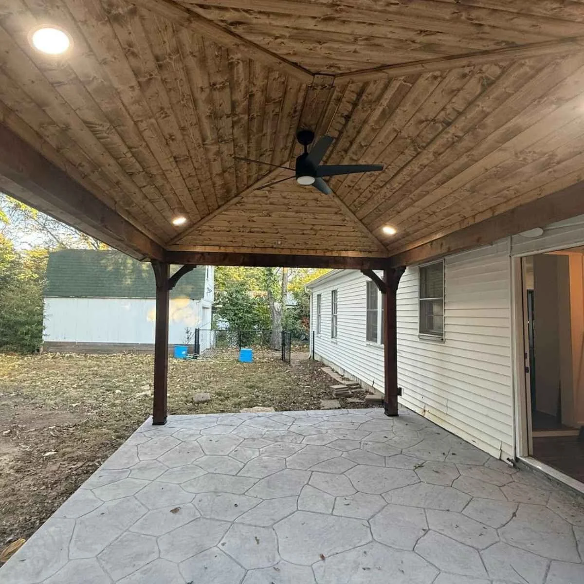 Covered backyard patio with wooden ceiling, ceiling fan, built-in lights, and concrete patterned floor, attached to a house.