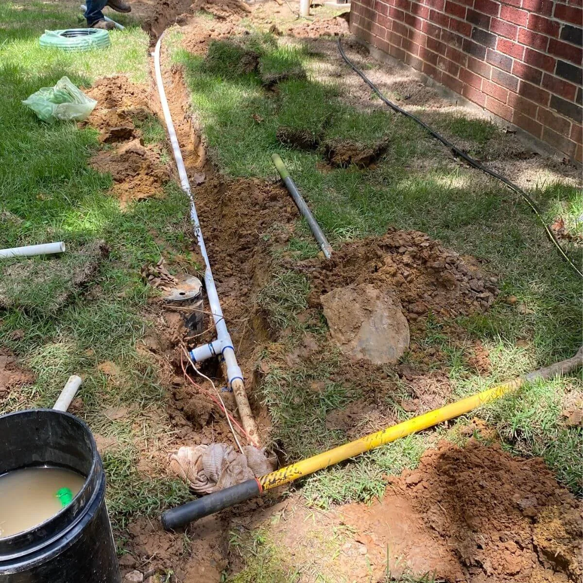 Installing underground sprinkler pipes outside a house with a brick wall, with some pipes already laid and others being installed, surrounded by dirt, grass, and construction tools.