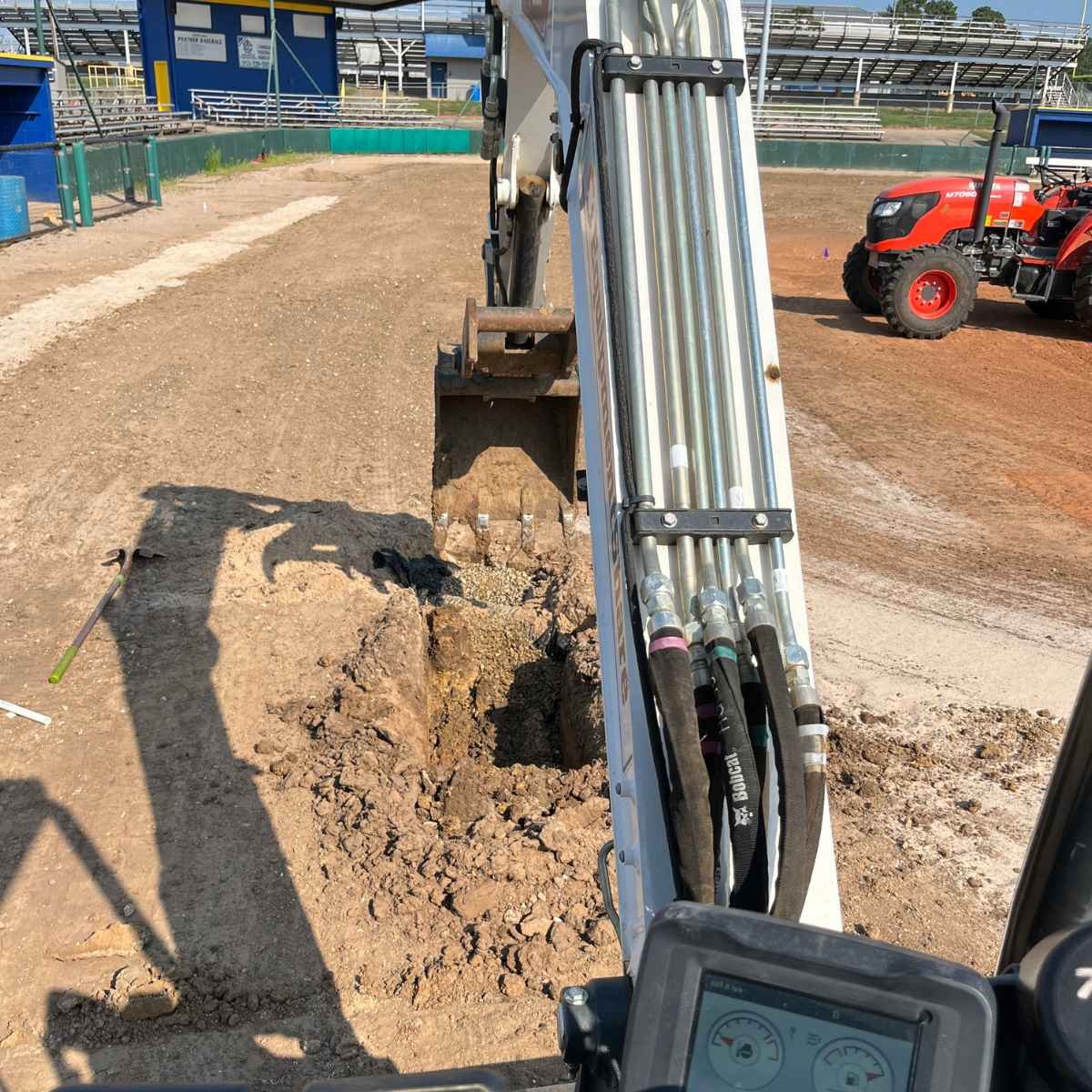 Construction site with dirt excavation, earth-moving machinery, and a red tractor in the background during an irrigation installation project on an athletic field in Paris, Texas.