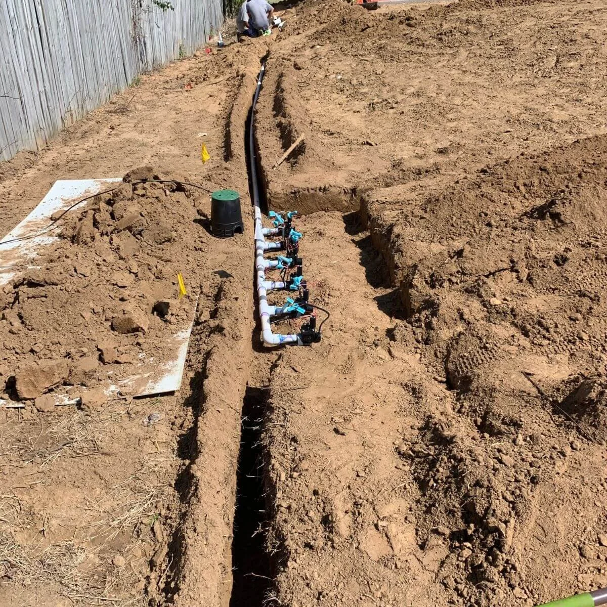 A trench in the ground with irrigation piping and valves installed, with a person working in the background.