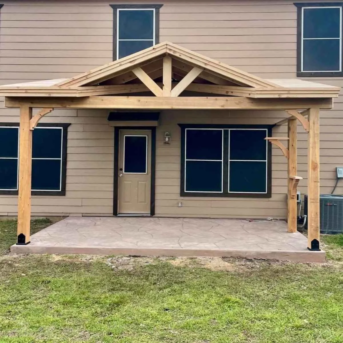 A house with a new wooden porch roof and support beams over a concrete patio. The house has beige siding, black window frames, and a beige door. There are three windows visible, two on the first floor and one on the second floor.